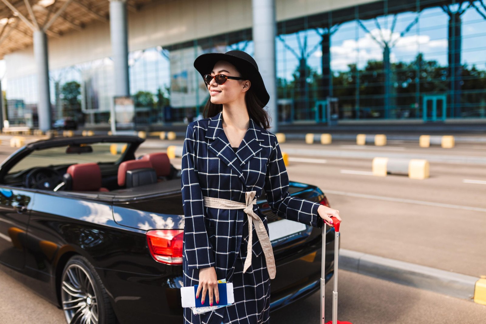beautiful-smiling-girl-sunglasses-black-hat-holding-passport-with-flight-ticket-joyfully-looking-aside-near-airport-with-cabriolet-car-background-min